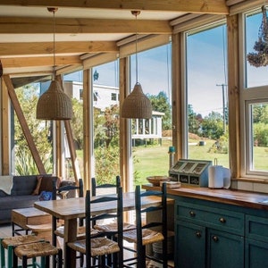 Puede incluir: Un comedor con una mesa y sillas de madera, un sof&aacute; azul y una vista de un campo de hierba a trav&eacute;s de grandes ventanas. Dos l&aacute;mparas colgantes de mimbre cuelgan sobre la mesa.