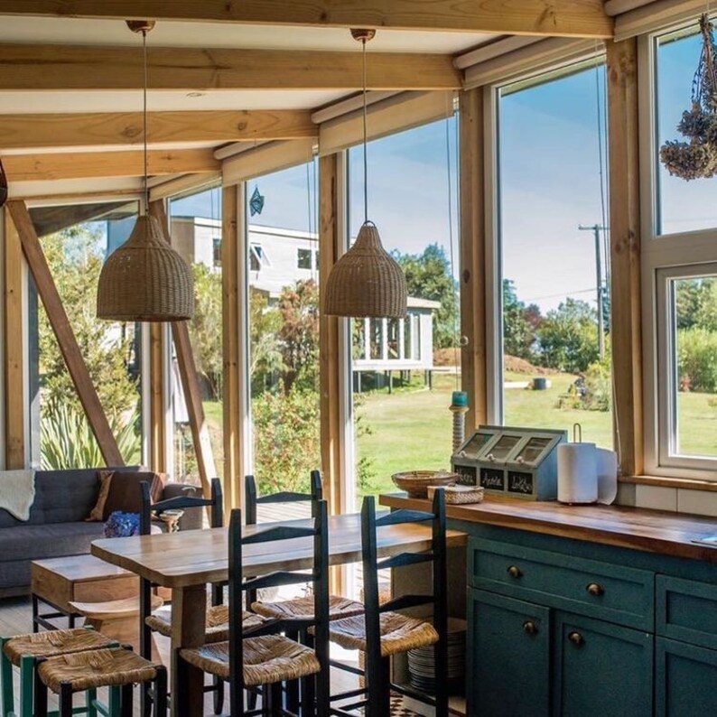 Puede incluir: Un comedor con una mesa y sillas de madera, un sof&aacute; azul y una vista de un campo de hierba a trav&eacute;s de grandes ventanas. Dos l&aacute;mparas colgantes de mimbre cuelgan sobre la mesa.