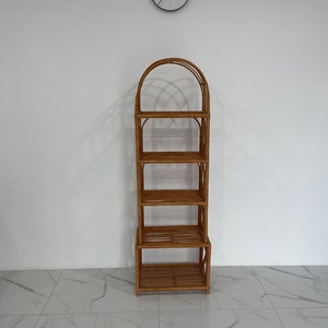 May include: A tall, natural woven rattan shelf unit with five shelves and an arched top. The shelf unit is standing on a white tiled floor.