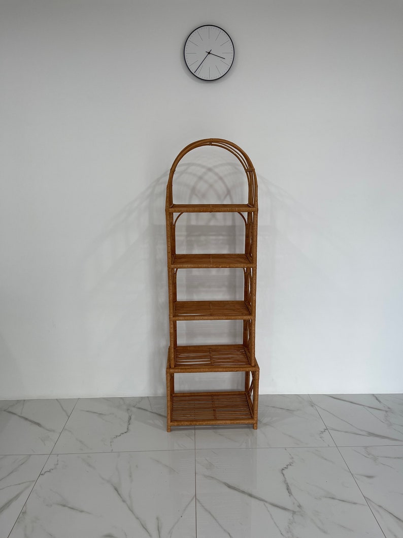 May include: A tall, natural woven rattan shelf unit with five shelves and an arched top. The shelf unit is standing on a white tiled floor.