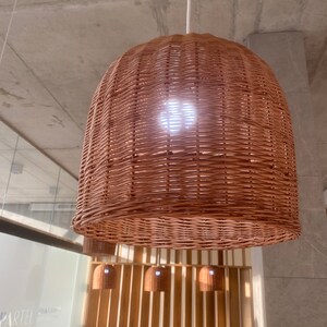 Puede incluir: Una gran l&aacute;mpara colgante redonda de mimbre tejido cuelga del techo. La l&aacute;mpara est&aacute; hecha de mimbre marr&oacute;n natural y tiene una superficie ligeramente texturizada. La l&aacute;mpara est&aacute; iluminada desde el interior, proyectando un c&aacute;lido resplandor.