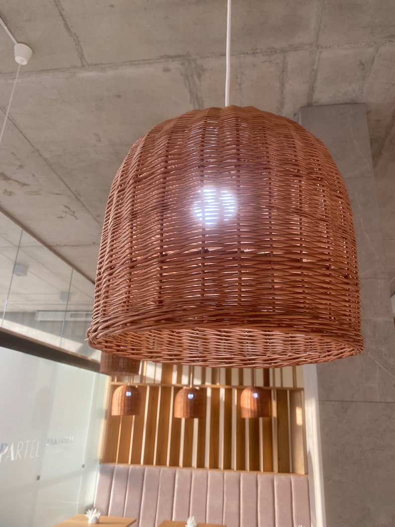 Puede incluir: Una gran l&aacute;mpara colgante redonda de mimbre tejido cuelga del techo. La l&aacute;mpara est&aacute; hecha de mimbre marr&oacute;n natural y tiene una superficie ligeramente texturizada. La l&aacute;mpara est&aacute; iluminada desde el interior, proyectando un c&aacute;lido resplandor.