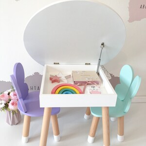 May include: A white children's table with a lift-top lid, two chairs, and a rainbow stacking toy. The chairs are purple and light blue with butterfly-shaped backs. The table has a white top and wooden legs.