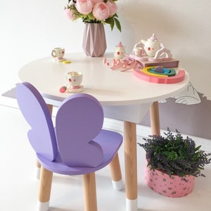May include: A white children's table and purple butterfly chair with wooden legs. The table is decorated with a pink flower arrangement, a tea set, and a rainbow toy.