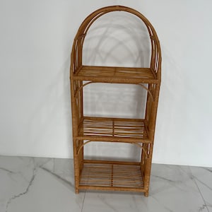 May include: A three-tiered natural wicker shelf unit with an arched top. The shelf is made of woven rattan and has a light brown color.