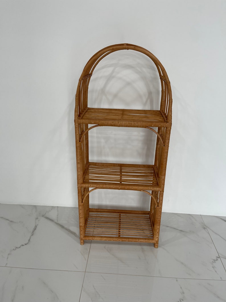 May include: A three-tiered natural wicker shelf unit with an arched top. The shelf is made of woven rattan and has a light brown color.