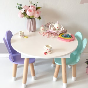 May include: A white oval table with two wooden chairs, one purple and one light blue. The chairs have white painted legs. A pink flower arrangement is in a vase on the table. There is a tea set and other toys on the table.