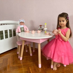 May include: A pink and white children's play table and chairs set with a doll and a young girl sitting at the table. The table has a white top and pink legs. The chairs are also pink and white. The doll is wearing a pink and white dress and a hat. The girl is wearing a pink dress.