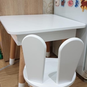 Op de afbeelding: Witte houten kindertafel en stoel set. De tafel heeft een vierkant blad en vier poten. De stoel heeft een ronde zitting en twee rugleuningen in de vorm van konijnenoren. De poten van zowel de tafel als de stoel zijn wit geverfd.