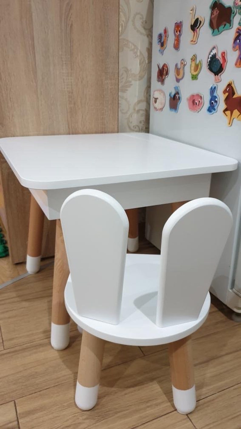 Op de afbeelding: Witte houten kindertafel en stoel set. De tafel heeft een vierkant blad en vier poten. De stoel heeft een ronde zitting en twee rugleuningen in de vorm van konijnenoren. De poten van zowel de tafel als de stoel zijn wit geverfd.