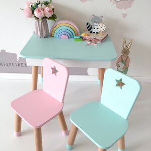 Op de afbeelding: Een lichtblauwe kindertafel met twee stoelen. E&eacute;n stoel is roze en de andere is lichtblauw. Beide stoelen hebben een stervormige uitsparing op de rugleuning. De tafel heeft een lichtblauw tafelblad en houten poten. Op de tafel staan speelgoed, waaronder een regenboog, een houten dier en een boek.
