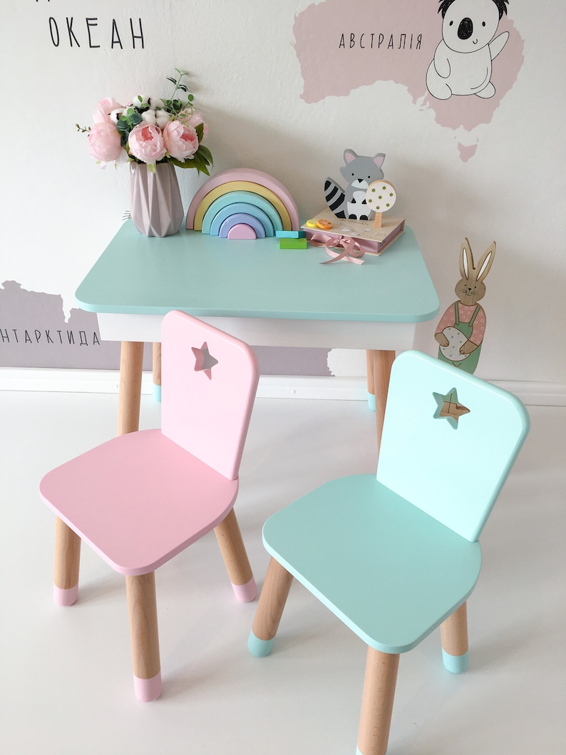 Op de afbeelding: Een lichtblauwe kindertafel met twee stoelen. E&eacute;n stoel is roze en de andere is lichtblauw. Beide stoelen hebben een stervormige uitsparing op de rugleuning. De tafel heeft een lichtblauw tafelblad en houten poten. Op de tafel staan speelgoed, waaronder een regenboog, een houten dier en een boek.