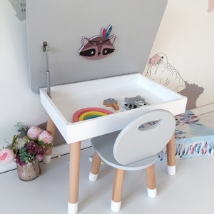 Op de afbeelding: Een witte en grijze kindertafel- en stoelsets met een opklapbaar deksel. De tafel heeft een houten onderstel met wit geverfde poten. De stoel heeft een grijze zitting en rugleuning met houten poten. Het tafelblad heeft een regenboog speelgoed en een wasbeer sticker.