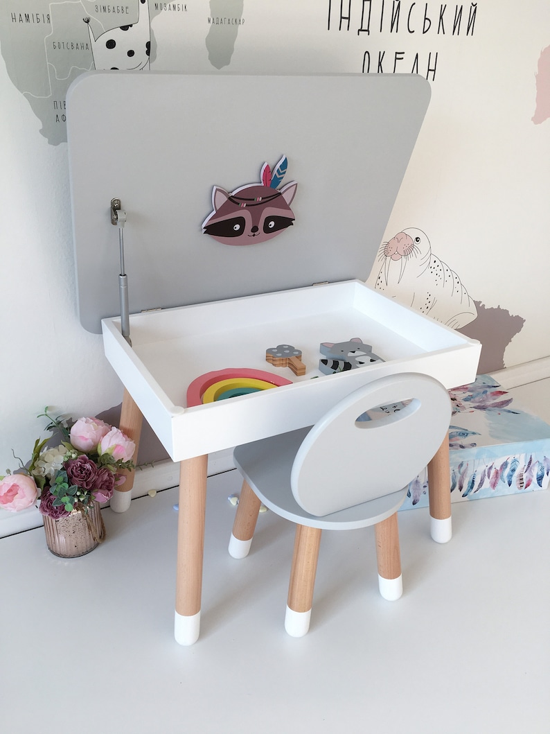Op de afbeelding: Een witte en grijze kindertafel- en stoelsets met een opklapbaar deksel. De tafel heeft een houten onderstel met wit geverfde poten. De stoel heeft een grijze zitting en rugleuning met houten poten. Het tafelblad heeft een regenboog speelgoed en een wasbeer sticker.
