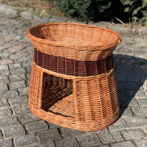 May include: A brown wicker pet bed with a raised oval top and a lower level with an opening. The bed has a brown woven band around the middle.