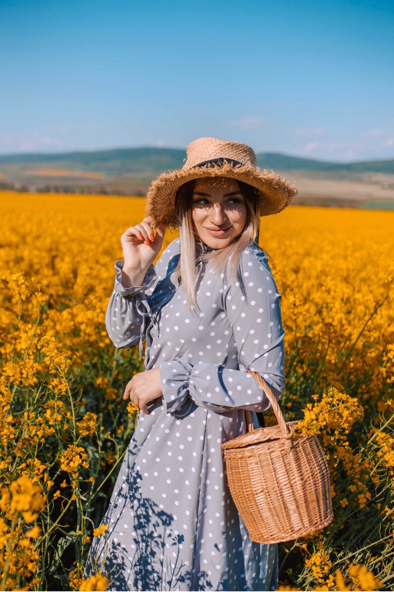 K&ouml;nnte beinhalten: Eine Frau in einem grauen Kleid mit wei&szlig;en Punkten und einem Strohhut steht in einem Feld mit gelben Blumen. Sie h&auml;lt einen Weidenkorb.