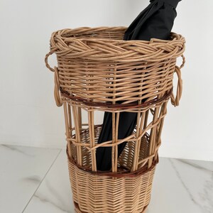 Pu&ograve; includere: Un portaombrelli in vimini intrecciato con un ombrello nero all'interno. Il portaombrelli ha una forma cilindrica con una fascia in vimini marrone intorno al centro.