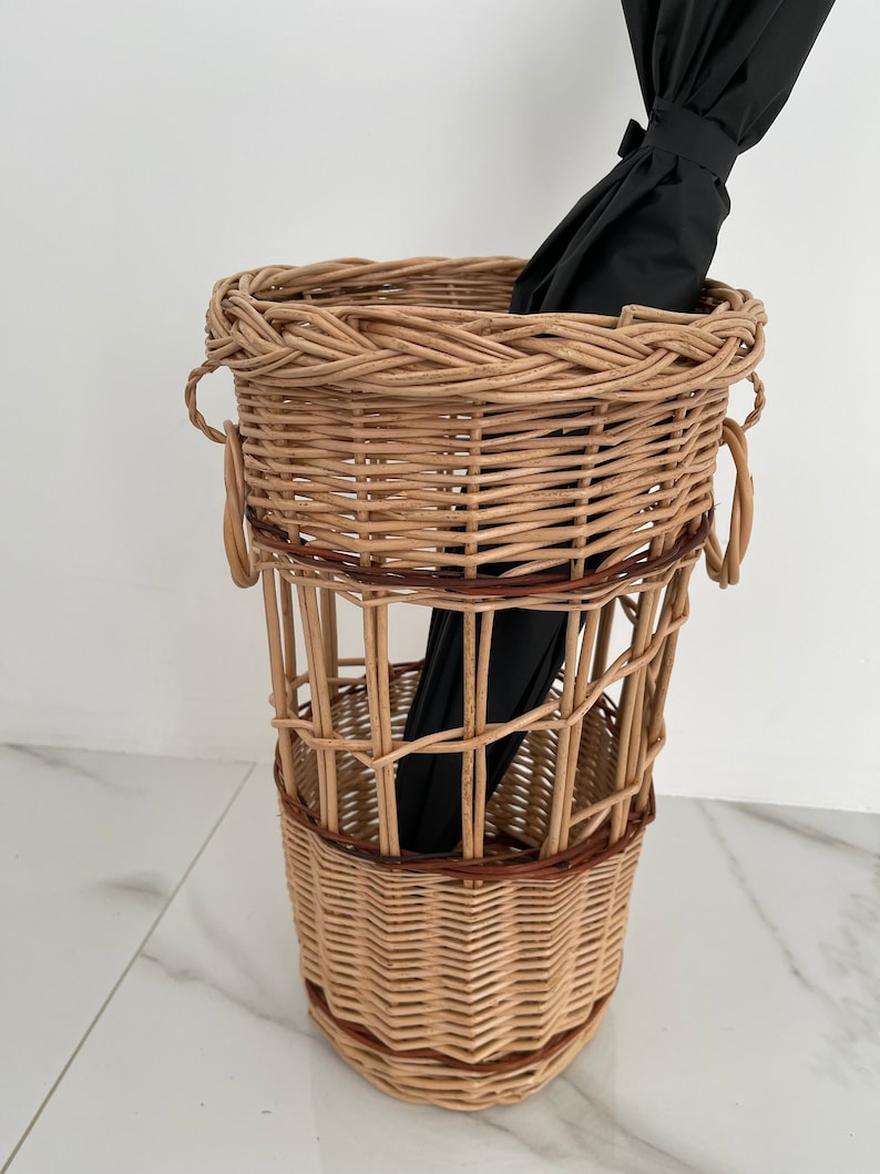 Pu&ograve; includere: Un portaombrelli in vimini intrecciato con un ombrello nero all'interno. Il portaombrelli ha una forma cilindrica con una fascia in vimini marrone intorno al centro.