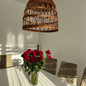 May include: A bouquet of red roses in a clear glass vase on a white table with a woven wicker pendant light hanging above. The table is set with brown velvet chairs.