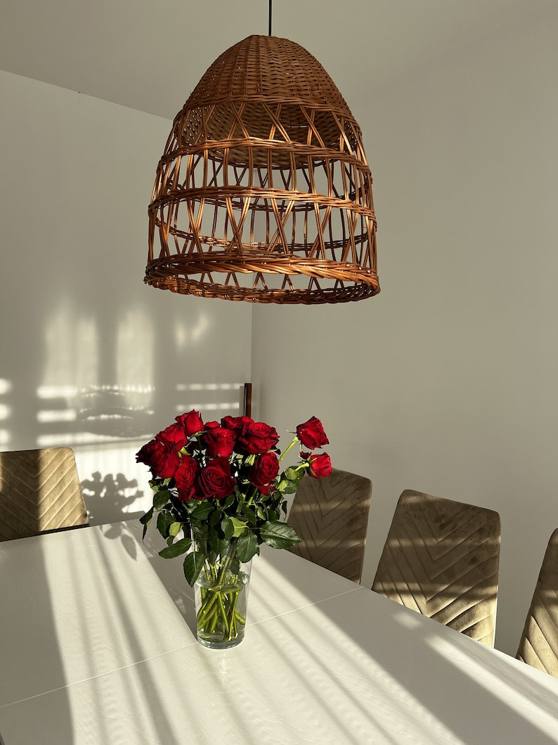 May include: A bouquet of red roses in a clear glass vase on a white table with a woven wicker pendant light hanging above. The table is set with brown velvet chairs.