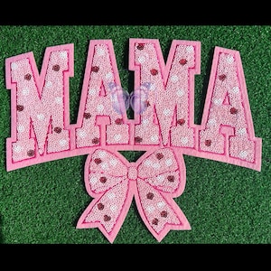 May include: A pink felt sign spelling "MAMA" in large, block letters. The letters are filled with pink sequins and white polka dots, with red heart accents. A pink bow is attached below the letters.