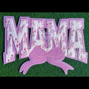 May include: A purple felt applique with the word "MAMA" in white letters with a pink floral pattern. A large pink bow is attached to the bottom of the applique.
