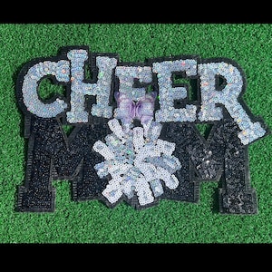 Peut inclure: Un motif "CHEER MOM" à paillettes argentées et noires, avec un papillon violet et un pompon argenté. Le motif est posé sur un fond vert, parfait pour les fans de cheerleading.