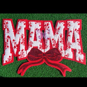 May include: A decorative sign spelling "MAMA" in red and white. The letters are outlined in red and filled with white fabric and red floral embroidery. A red sequined bow is at the bottom.