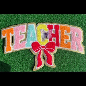 Puede incluir: Aplicación de fieltro colorido que deletrea "TEACHER" en naranja, rosa, turquesa, amarillo y rojo, delineada con purpurina dorada. Un gran lazo rojo está centrado debajo de la palabra, también delineado con purpurina dorada. La aplicación está sobre un fondo verde.