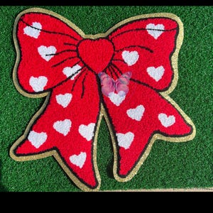 May include: A large, red bow-shaped rug with white heart patterns. The rug has a gold glitter border and a red heart in the center. A pink and purple butterfly is placed on the bow. The rug is on a green surface.