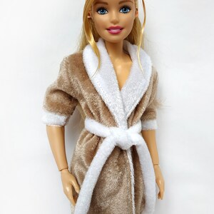 May include: A brown plush robe with a white trim and a white sash. The robe has a shawl collar and is worn by a doll with blonde hair and blue eyes.