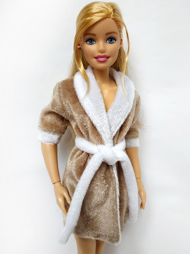 May include: A brown plush robe with a white trim and a white sash. The robe has a shawl collar and is worn by a doll with blonde hair and blue eyes.