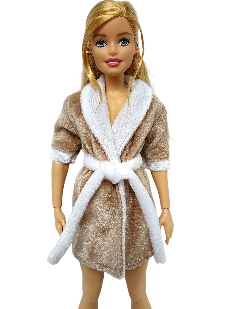 May include: A brown plush robe with a white trim and a white tie belt. The robe is worn by a doll with blonde hair and blue eyes.
