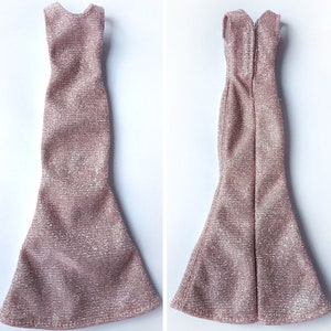 May include: Two doll-sized, sleeveless, rose-gold dresses. The dresses have a textured, shimmering fabric. One dress is shown from the front, the other from the back, revealing a zipper. Both dresses have a mermaid-style skirt.
