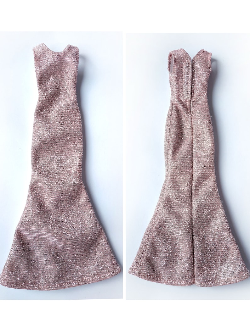 May include: Two doll-sized, sleeveless, rose-gold dresses. The dresses have a textured, shimmering fabric. One dress is shown from the front, the other from the back, revealing a zipper. Both dresses have a mermaid-style skirt.