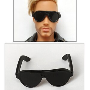 K&ouml;nnte beinhalten: Schwarze Plastik-Sonnenbrille mit abgerundeter Aviator-Form. Die Sonnenbrille ist f&uuml;r eine Puppe oder eine Actionfigur konzipiert.