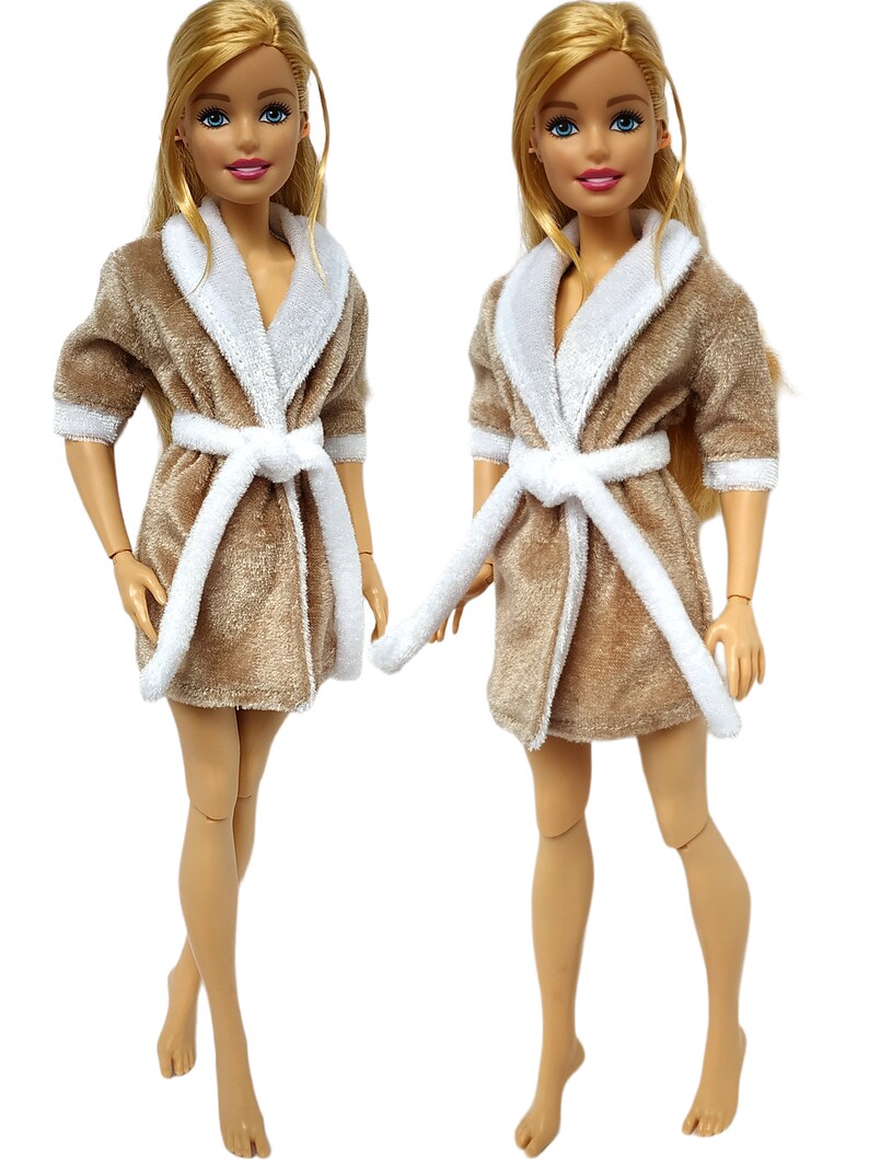 May include: Two blonde dolls wearing beige plush bathrobes with white trim and white sashes. The robes have long sleeves and a shawl collar.