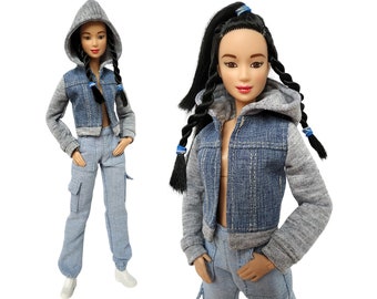 May include: A doll with long black hair styled in braids with blue hair ties. The doll is wearing a blue denim jacket with grey sleeves, a grey hooded sweatshirt, and blue denim pants.