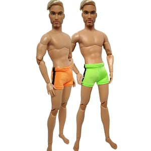 May include: Two articulated male dolls wearing swim trunks. One doll is wearing orange swim trunks with black trim. The other doll is wearing green swim trunks with black trim.