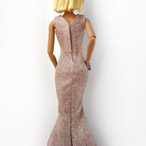 May include: A doll wearing a shimmering, rose-gold evening gown with a mermaid silhouette. The dress features a V-neckline and a zipper down the back. The doll has short, blonde hair.