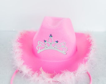 May include: A bright pink cowboy hat featuring a silver-toned tiara embellishment on the front. The tiara is decorated with small, colorful faux gemstones. The brim of the hat is trimmed with light pink feathers and silver metallic tinsel.