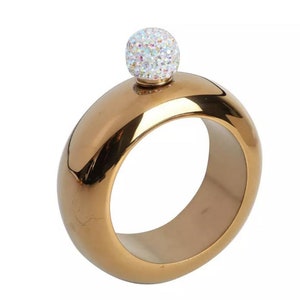 May include: A circular gold-toned metal flask with a smooth, reflective surface and a central opening. The flask features a spherical screw-top cap covered in small, iridescent, multi-colored crystals that catch the light with a shimmering effect.