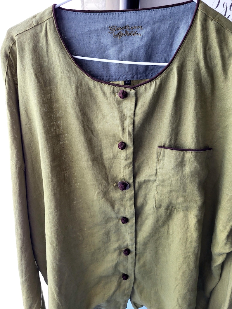 Puede incluir: Una camisa de lino verde con ribete marr&oacute;n y cierre de botones en la parte delantera. La camisa tiene un bolsillo en el lado izquierdo y una etiqueta que dice "Kuskoon Apparel" en el interior del cuello.