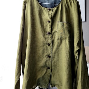 Puede incluir: Una camisa de lino verde con cierre de botones marrones y un solo bolsillo en el lado izquierdo. La camisa tiene un cuello redondo y mangas largas.