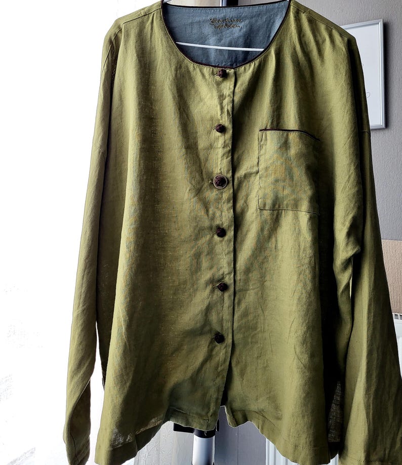 Puede incluir: Una camisa de lino verde con cierre de botones marrones y un solo bolsillo en el lado izquierdo. La camisa tiene un cuello redondo y mangas largas.