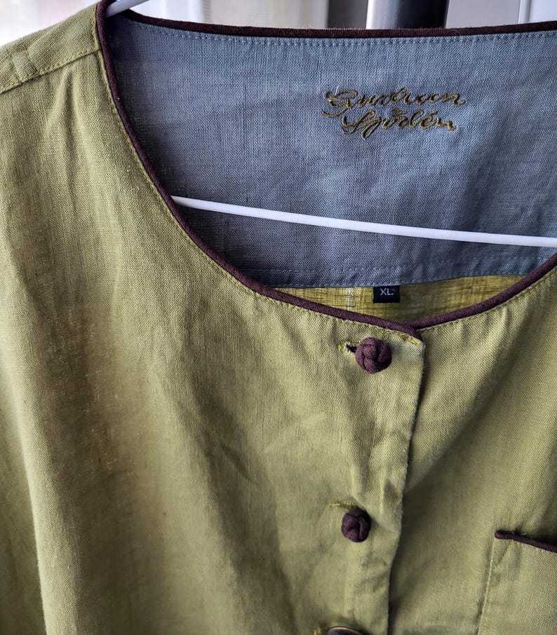 Puede incluir: Una camisa de lino verde con ribetes y botones marrones. La camisa tiene un cuello mandar&iacute;n y una etiqueta que dice "Runaway Sprinkles" y "XL".