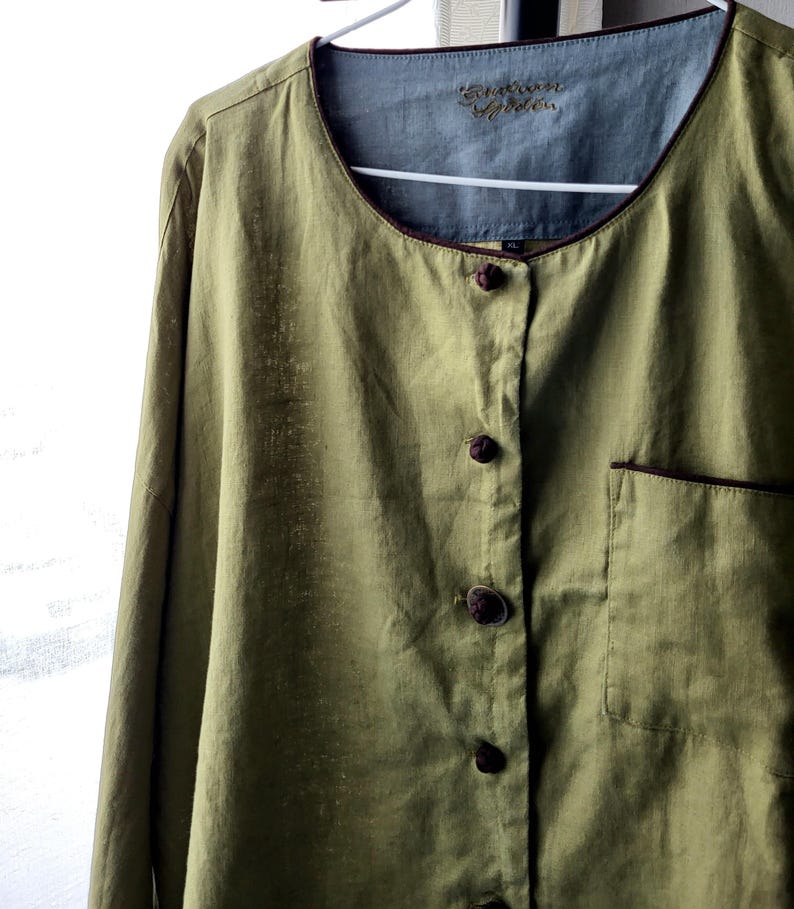 Puede incluir: Una camisa de lino verde con ribete marr&oacute;n alrededor del cuello y los bolsillos. La camisa tiene un frente abotonado con botones anudados. La etiqueta de la camisa dice "Embroidered Garden".