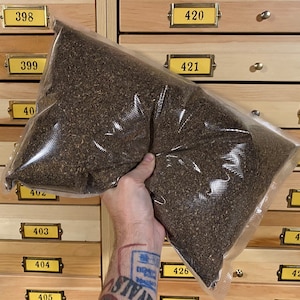 Peut inclure: Un grand sac en plastique transparent rempli d'herbes séchées brunes en vrac. Le sac est tenu par une main avec un tatouage. Le sac est devant un meuble en bois avec des tiroirs numérotés.