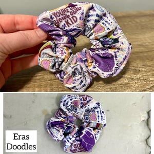 May include: Purple and white scrunchie with a variety of colorful stickers and text, including "You're doing your own kid" and "Eras Doodles".