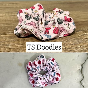 May include: Pink fabric scrunchie with a pattern of red hearts, stars, and other small designs. The scrunchie is made of a soft, textured fabric and is tied in a knot.
